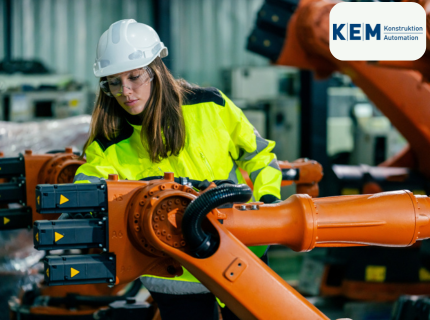Eine Person in einer gelben Sicherheitsjacke und einem weißen Helm steht neben einem orangefarbenen Industrieroboterarm mit grauen Steuerboxen.