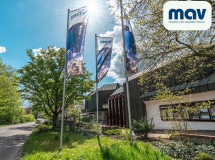 Three flags featuring the 'mav' logo are waving in front of a modern building on a sunny day with a blue sky and trees.